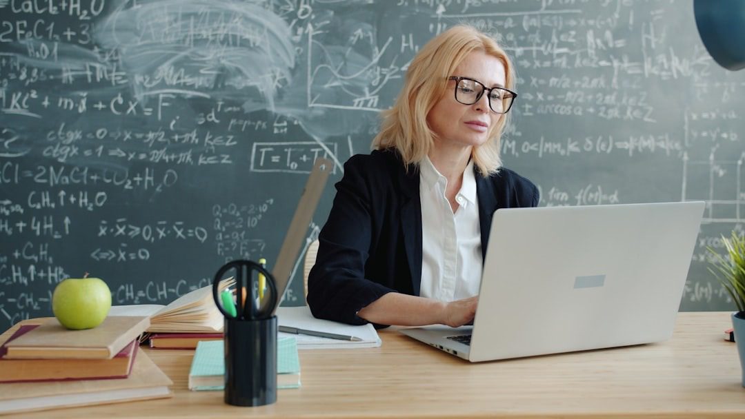 Woman works on laptop at desk with math equations on chalkboard.